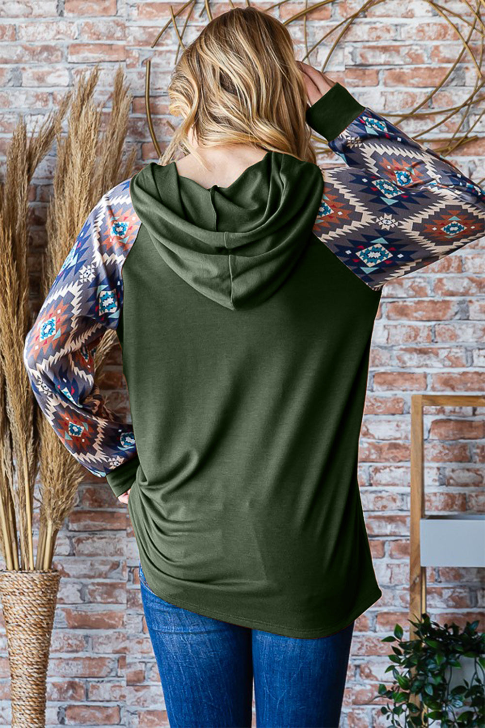 A woman poses indoors wearing a Heimish Full Size Half Button Printed Long Sleeve Hooded Top, showcasing its unique sleeve print design. Holding the hood with one hand, she complements her casual and on-trend outfit with blue jeans. The background features brick walls and tall dried plants.