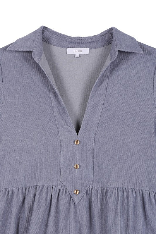 The woman in the Corduroy tiered dress, featuring a deep V-neck, smiles as she gently touches her forehead, showcasing the light blue fabric cascading elegantly.