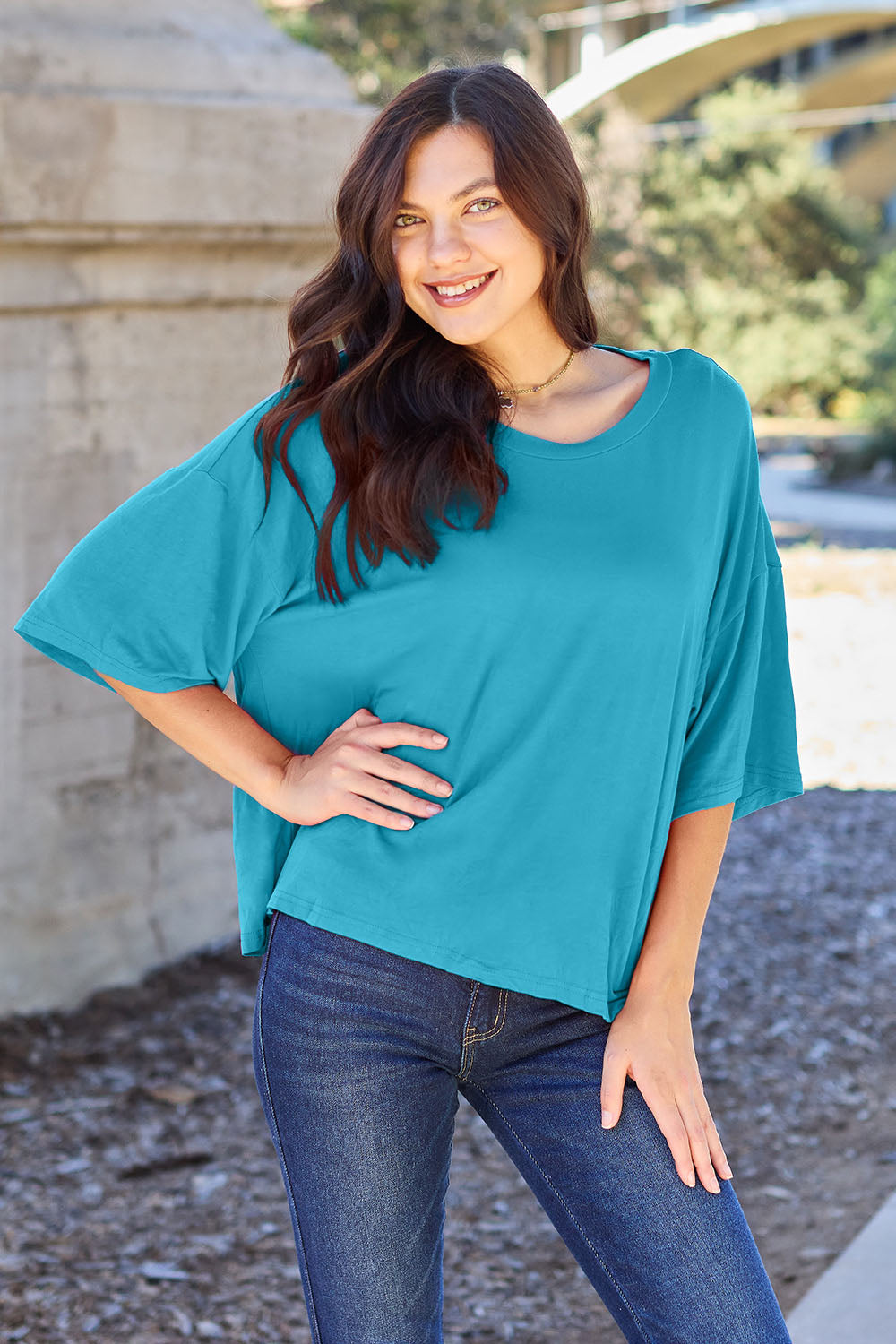 A person with long dark hair and a bright smile stands outdoors, showcasing their basic style in a loose, slightly stretchy *Basic Bae Full Size Round Neck Drop Shoulder T-Shirt* in pink and blue jeans, with one hand on their hip.