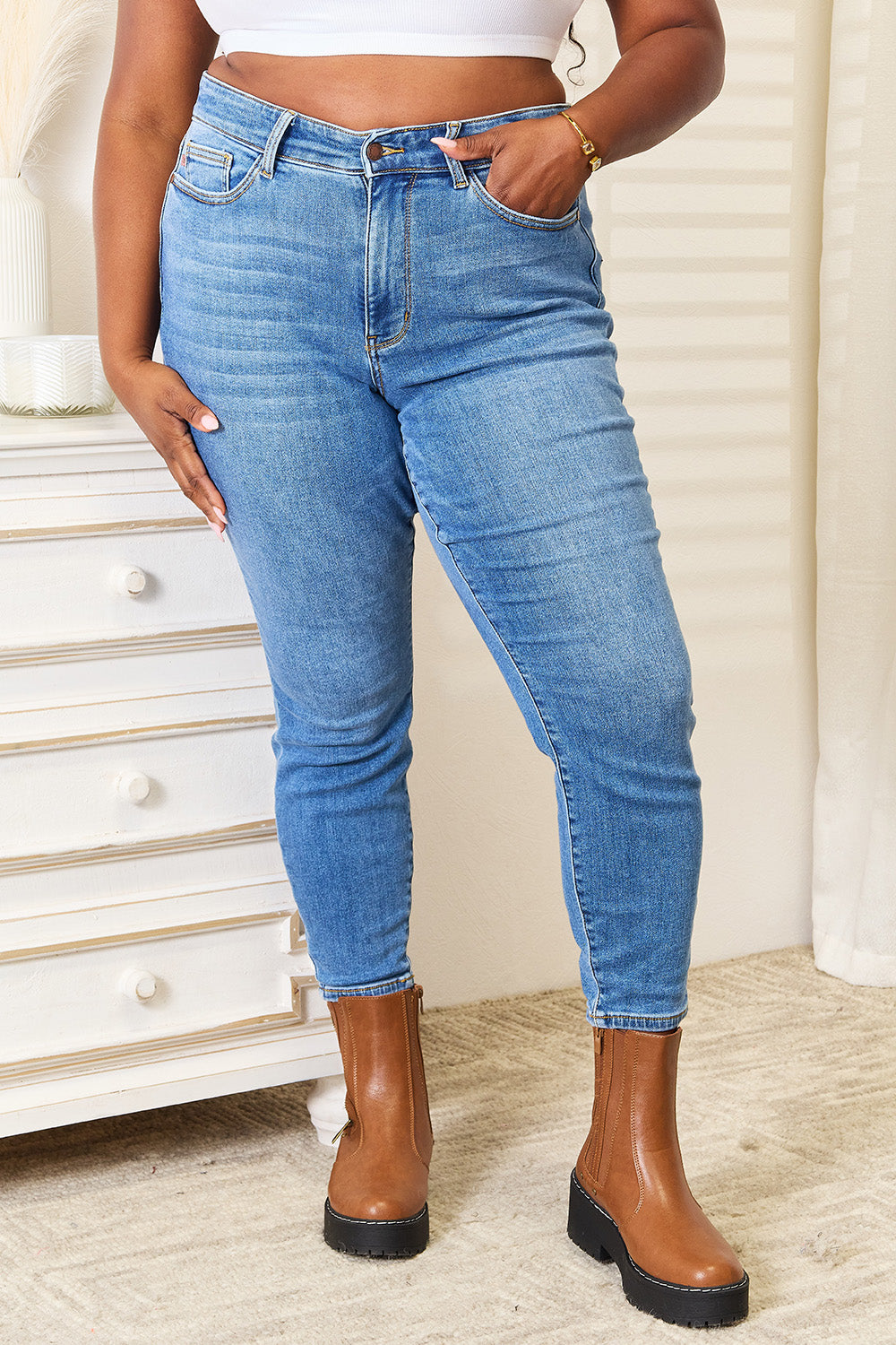 A person wearing Judy Blue Full Size High Waist Skinny Jeans in medium ash wash with a flattering fit, a white top, and brown boots stands near a white dresser, casually slipping one hand into their pocket.