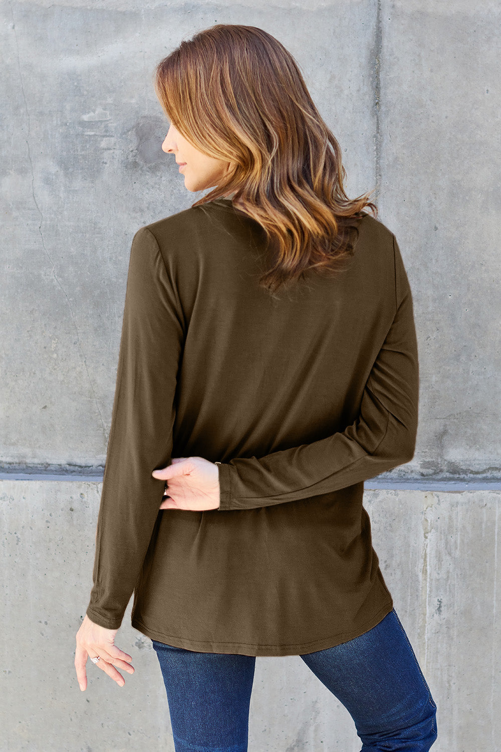 A woman with light brown hair stands against a concrete wall, wearing the Basic Bae Full Size V-Neck Long Sleeve Top in olive green and blue jeans, with a white backpack on her shoulder. Her outfit exudes basic style and is made from polyester, ensuring it’s both comfortable and suitable for machine wash cold. She is looking at the camera.