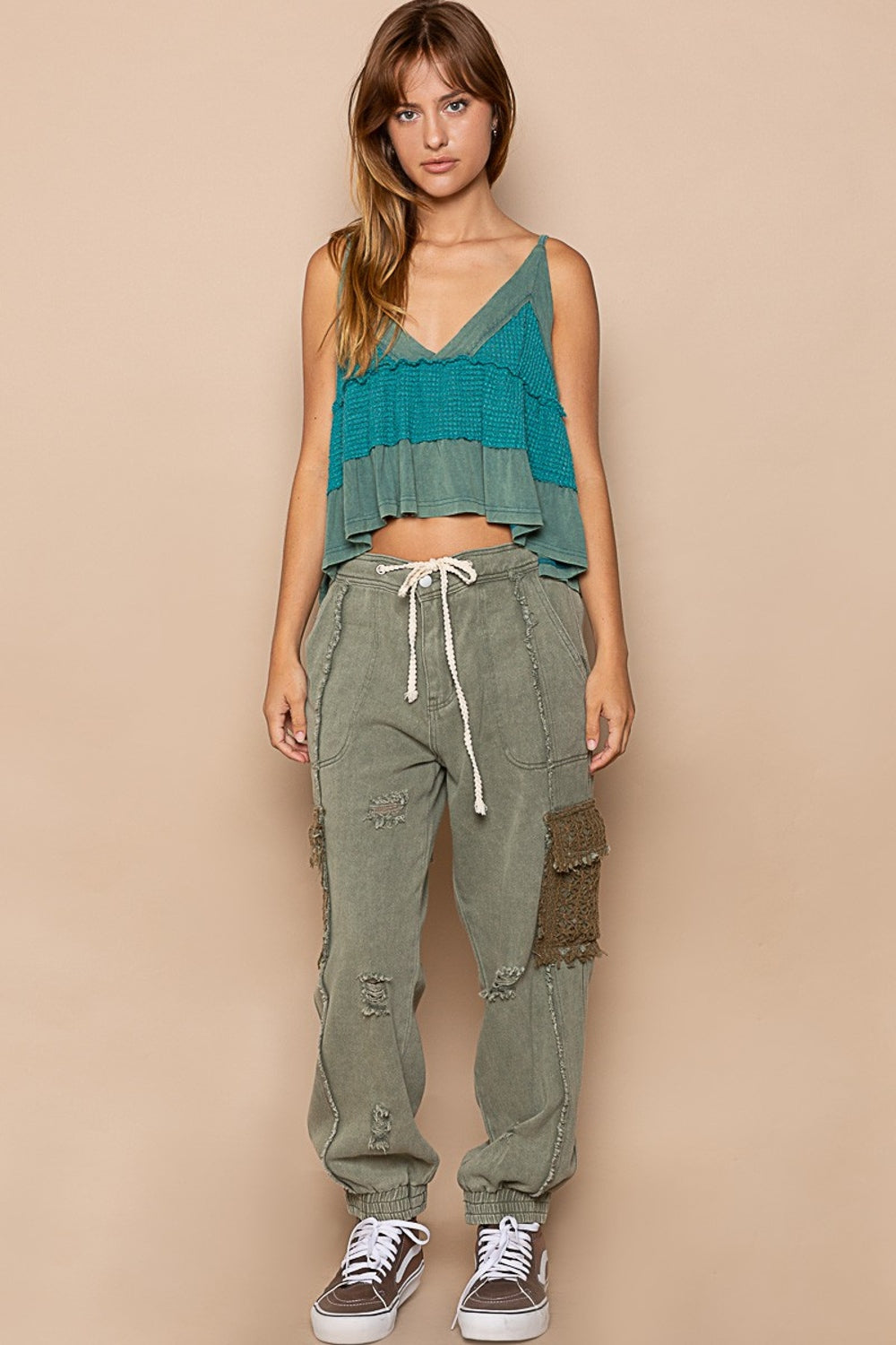 A person sporting POL Distressed Cargo Denim Joggers with Crochet Pockets in green and a white top stands casually on a colorful rug, hands tucked in pockets.