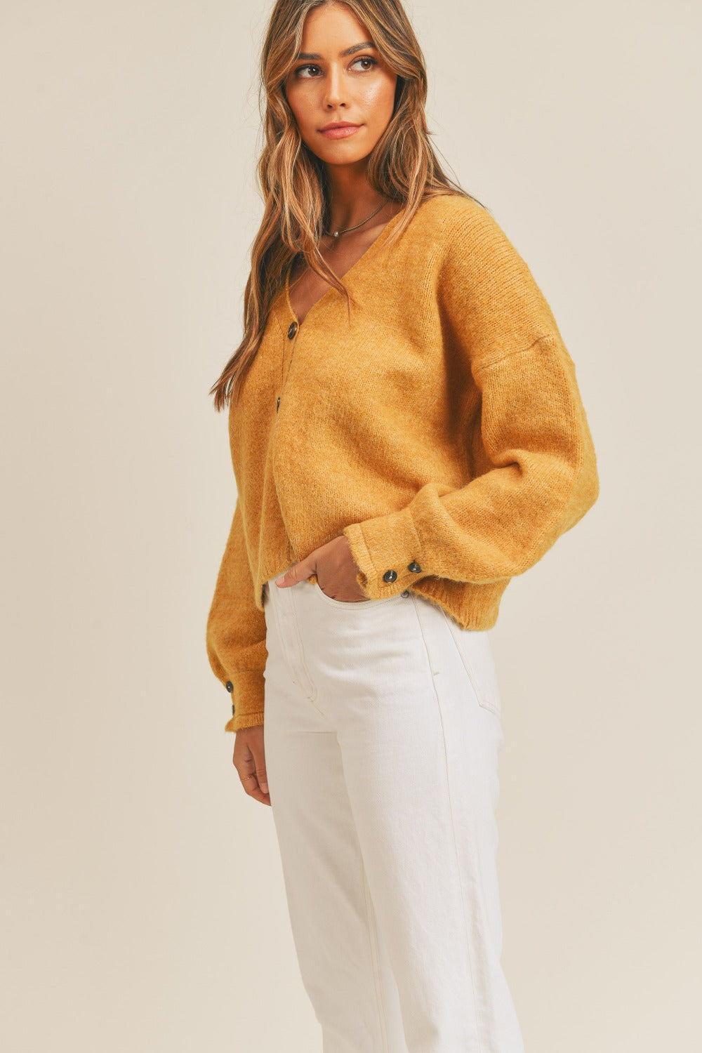 A person wears the MABLE Long Sleeve Button Down Sweater Cardigan in mustard, paired with white high-waisted pants, standing in front of a plain background.