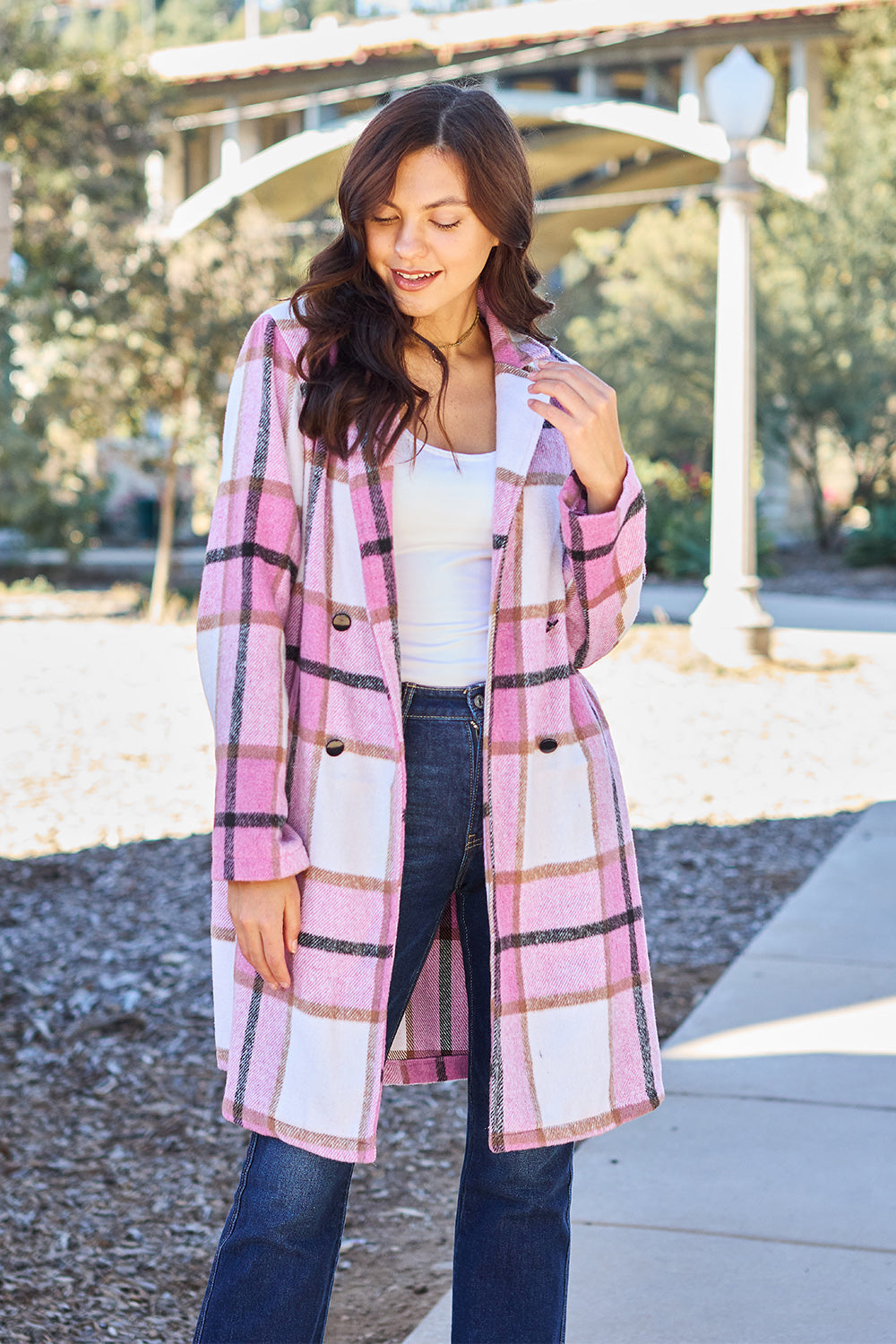 A woman stands on a sidewalk wearing the Double Take Full Size Plaid Button Up Lapel Collar Coat in pink and white over a white top and blue jeans, with her right hand lightly touching her hair. The polyester fabric of the coat makes it durable and perfect for an easy machine wash cold.