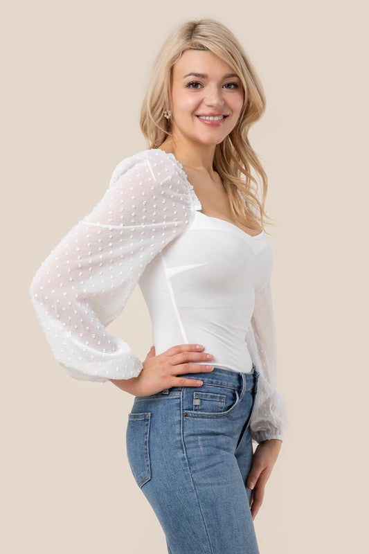 A person wearing the LS shirring sleeve bodysuit with Swiss dot mesh, featuring sheer, long puffed sleeves and a sweetheart neck, paired with light-colored jeans, stands against a plain background.