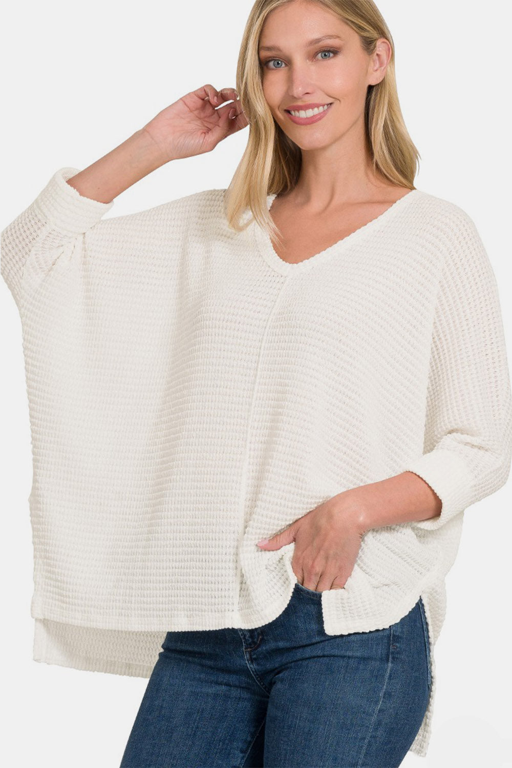 A smiling woman confidently poses in the Zenana Full Size Round Neck High-Low Slit Knit Top, which features a loose fit and three-quarter sleeves. She pairs the white knit top with blue jeans, striking a pose with one hand on her head and the other in her pocket.