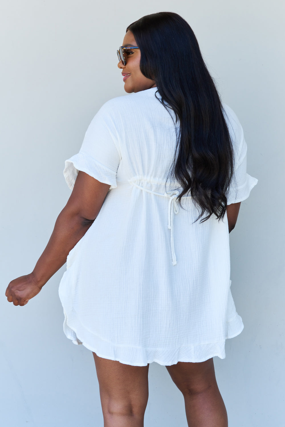 A person with long dark hair smiles while posing in the Ninexis Out Of Time Full Size Ruffle Hem Dress with Drawstring Waistband in White against a plain background.