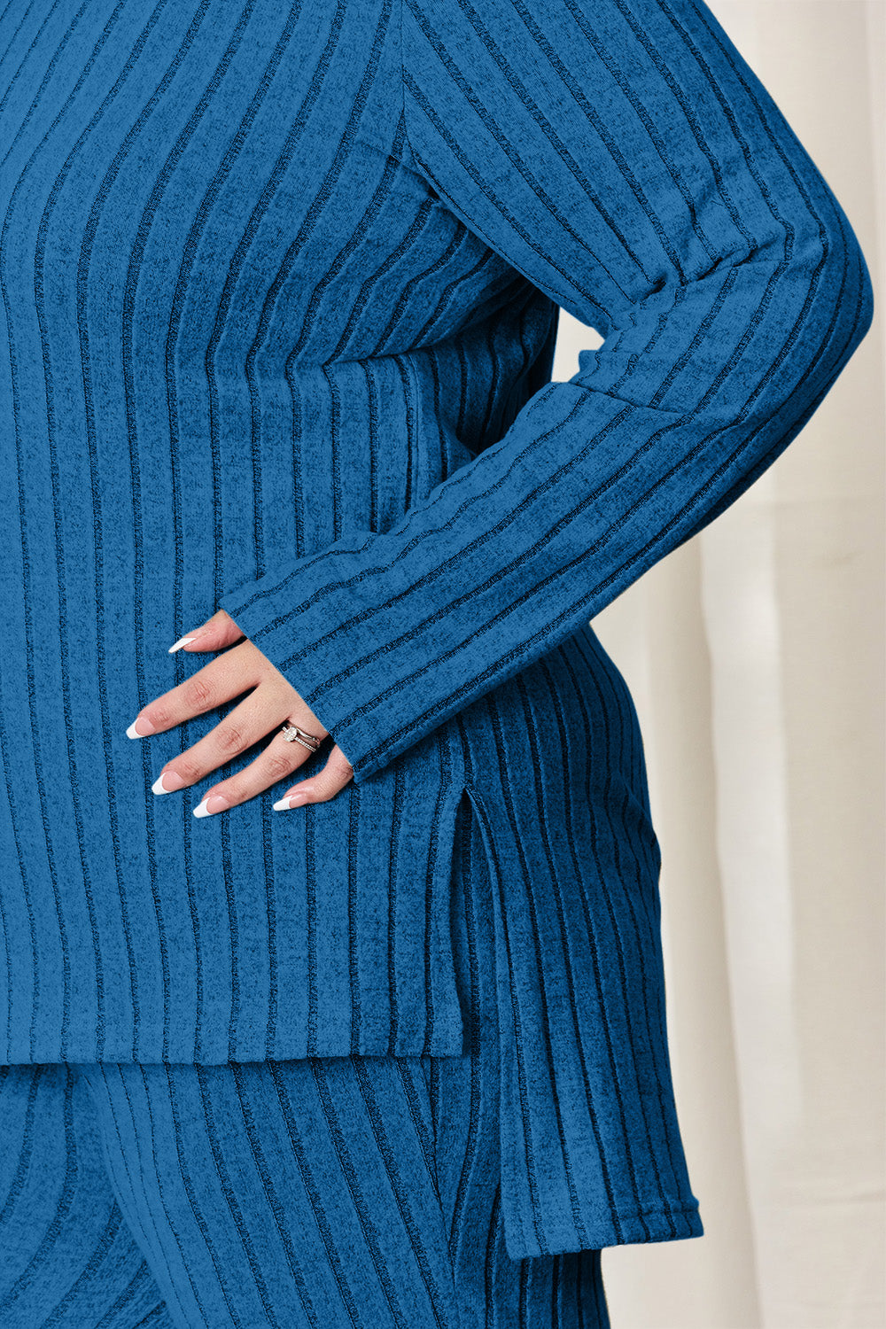 A person wearing the Basic Bae Full Size Ribbed High-Low Top and Wide Leg Pants Set in light gray, made from stretchy material, stands indoors in front of a white curtain.