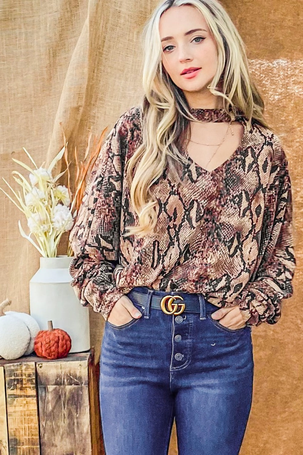 A woman wearing the And The Why Choker Neck Dolman Sleeve Snake Print Top paired with high-waisted jeans, smiling and posing in front of a brown background with decorative elements.