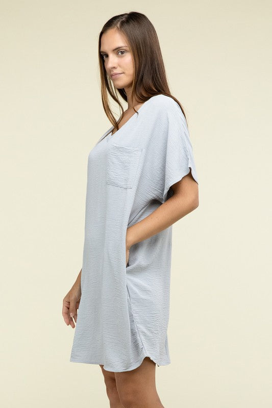 A woman wearing a loose light brown Woven Airflow V Neck T-Shirt Dress with Pockets stands against a plain background. Made from breathable fabric, this summer dress includes pockets where her hands rest as she smiles slightly.