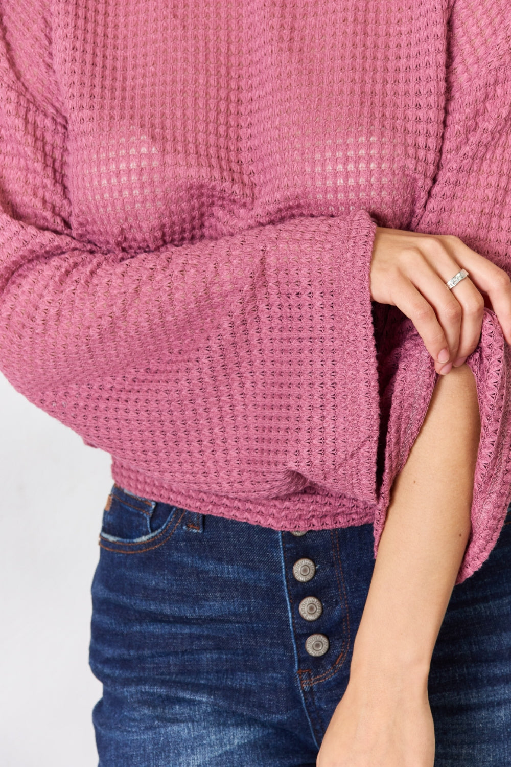 A person wearing a Culture Code Full Size Waffle-Knit Round Neck Long Sleeve Blouse in pink and jeans stands in front of a light background, complemented by a leafy plant on the left.