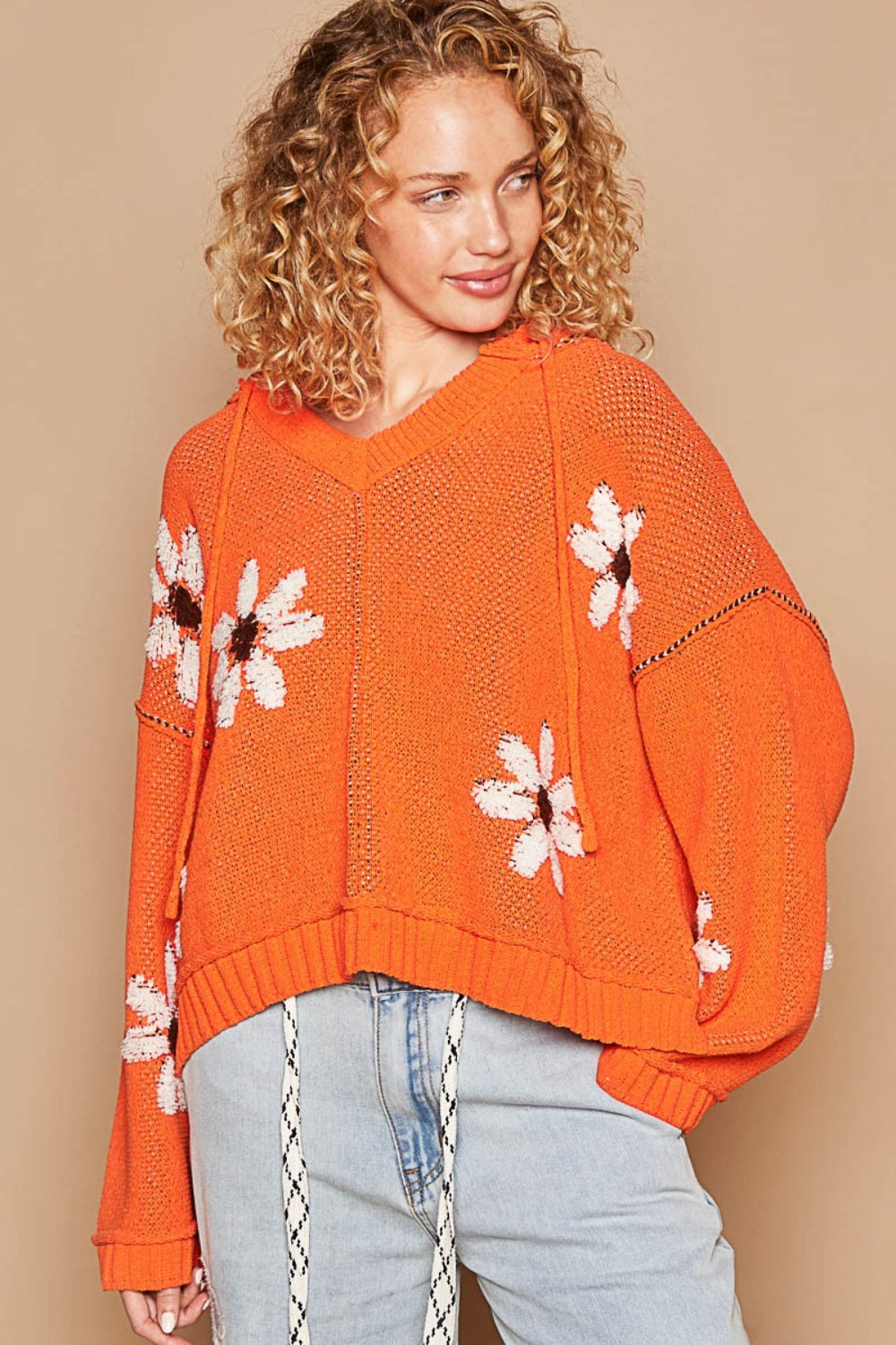 A person with curly blond hair is wearing the POL Floral Pattern Hooded High-Low Sweater in orange, featuring white and brown flower patterns against a solid beige background, paired with light blue jeans.