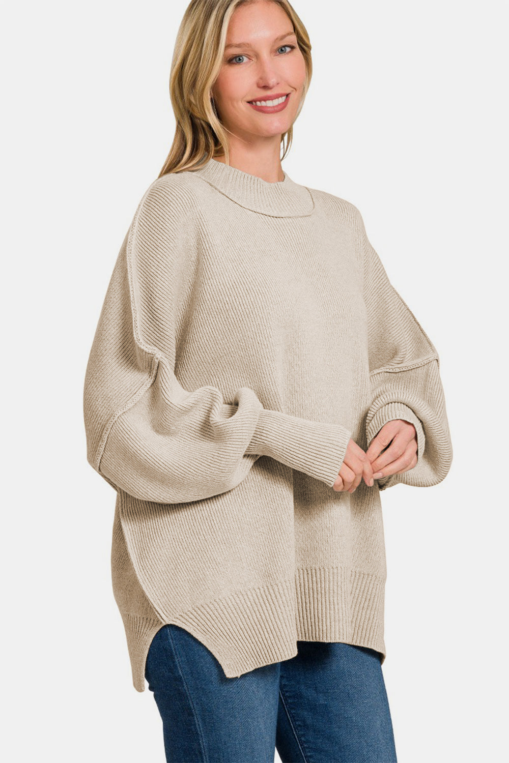 A person wearing the Zenana Side Slit Oversize Sweater, a cozy knit in beige, paired with jeans, smiling with one hand touching their hair.