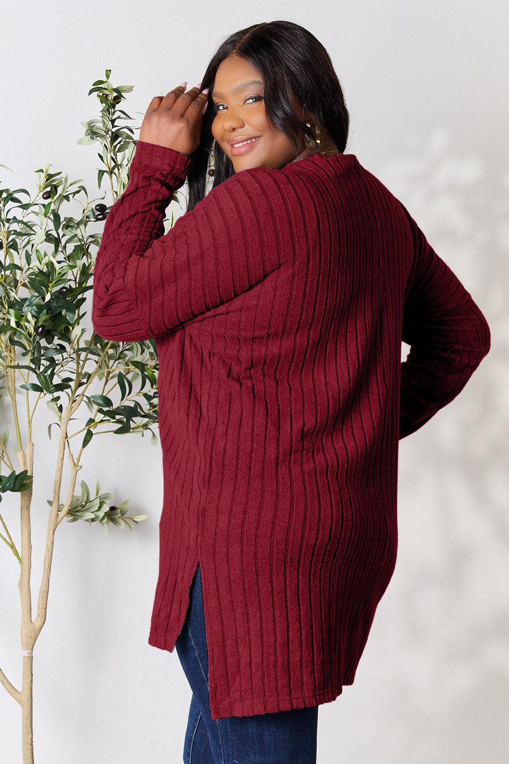 A person with shoulder-length hair smiles while wearing a beige ribbed sweater called the Basic Bae Full Size Ribbed Round Neck Long Sleeve Slit Top, along with matching beige pants, showcasing a basic style. They stand next to a green plant.