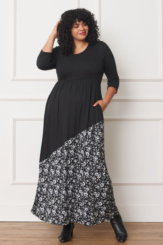 A woman with curly hair is wearing the Asymmetrical Leopard Accent Maxi Dress, featuring a long black design with a floral patterned section at the bottom. She stands against a white paneled wall, her hands tucked in her dress pockets, showcasing its elegant design.