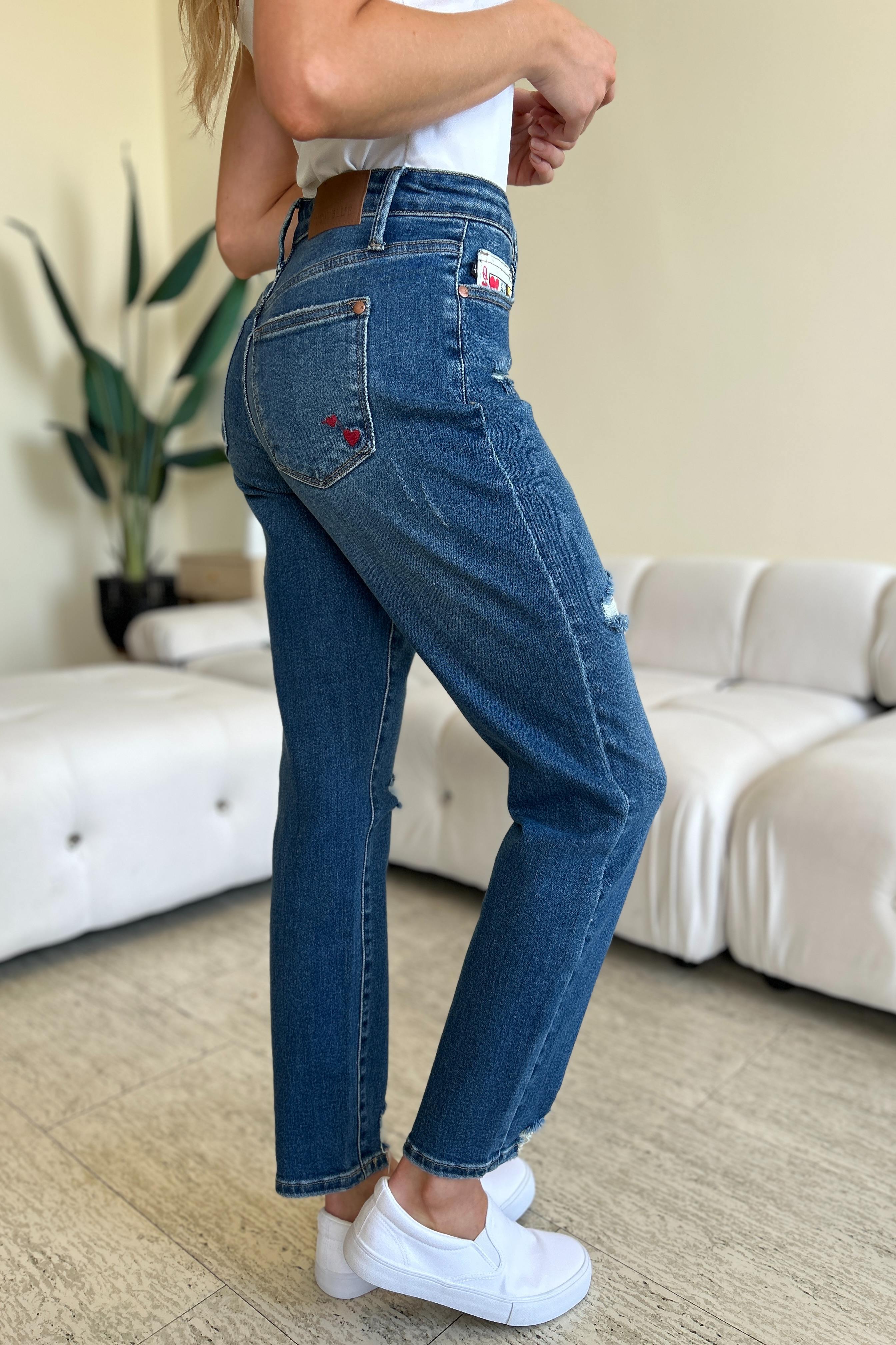 Two women stand side by side in a living room, both wearing stylish jeans and tank tops. One woman is pointing at the distinctive back pocket of her Judy Blue Full Size Queen Of Hearts Coin Pocket BF Jeans, while the other smiles.