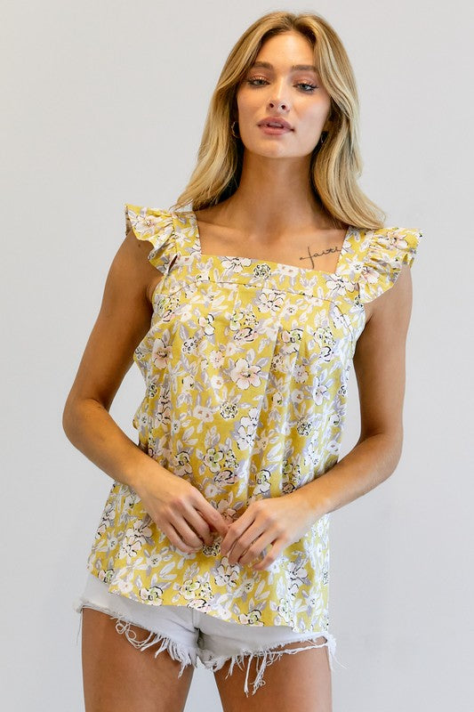 A woman in a vibrant summer wardrobe stands against a plain background, wearing the Floral Printed Ruffle Sleeveless Top paired with white shorts, with one hand casually tucked in her pocket.