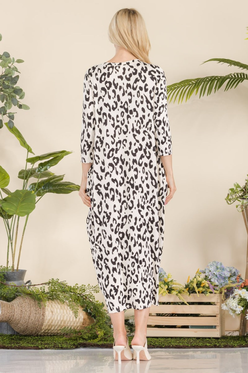 A woman exudes elegance in a beautifully decorated area filled with green plants and flowers, flaunting the Celeste Full Size Leopard Contrast Dress with Pockets, paired with white heels—an adaptable outfit ideal for any occasion.