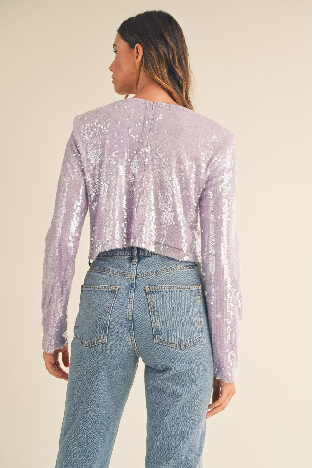 A woman with long, wavy hair wears a trendy MABLE Shoulder Padded Sequin Crop Top in light purple along with light blue jeans, standing against a plain off-white background.