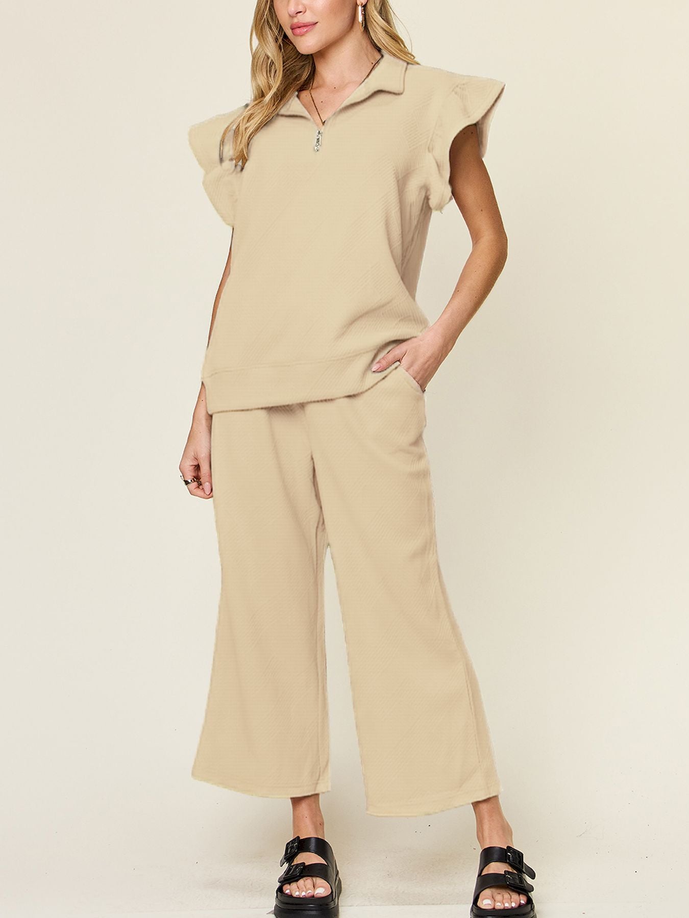 A woman stands wearing the Double Take Texture Ruffle Short Sleeve Top and Drawstring Wide Leg Pants Set in a slightly stretchy, light gray fabric with zipper detail. The two-piece outfit is complemented by black sandals, and she has one hand in her pocket. Machine wash cold for easy care.