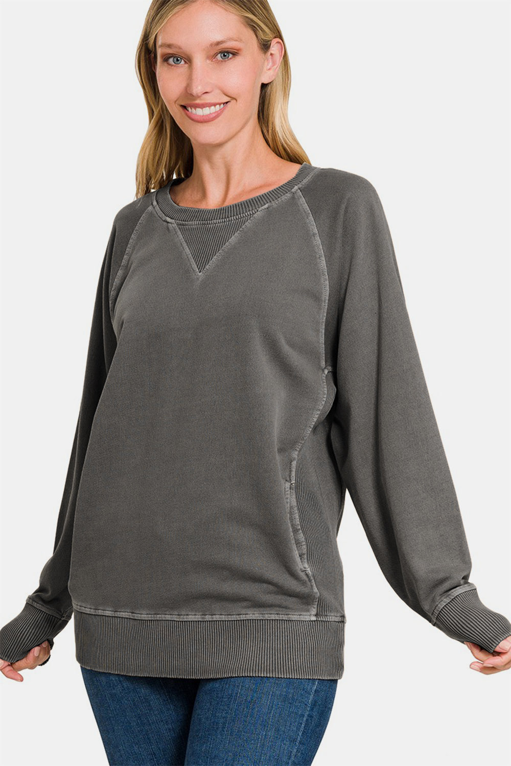 A person stands wearing a Zenana Round Neck Long Sleeve Sweatshirt, featuring a loose fit, dark grey color with front pockets and light stitching, paired with blue jeans. The 100% cotton fabric ensures comfort and durability.