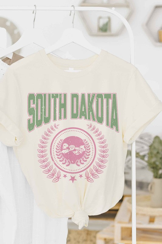 A person wearing a white SOUTH DAKOTA Graphic Tee, adorned with green and pink lettering, along with matching white shorts, stands indoors. The unisex sizing 100% cotton tee adds a comfortable yet stylish touch to their outfit.