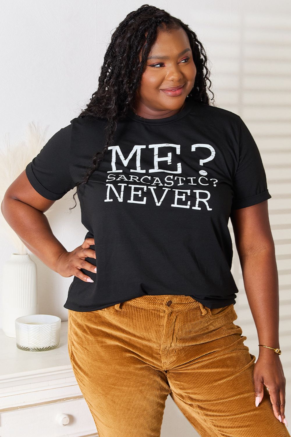 A woman with long dark hair smiles while wearing a trendy black Simply Love Letter Graphic Round Neck T-Shirt featuring the text "ME? Sarcastic? NEVER" and brown corduroy pants. She stands in a room with white walls and decor.