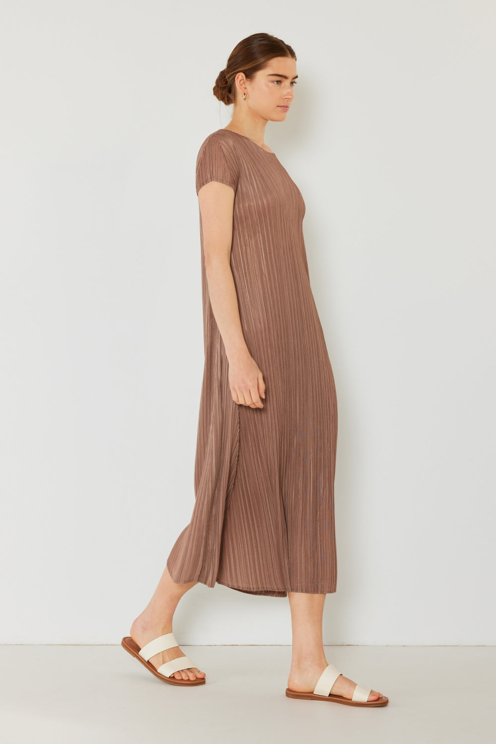 A person stands against a light wall, wearing an elegant Marina West Swim Pleated Cap Sleeve A-Line Dress in black and white sandals.