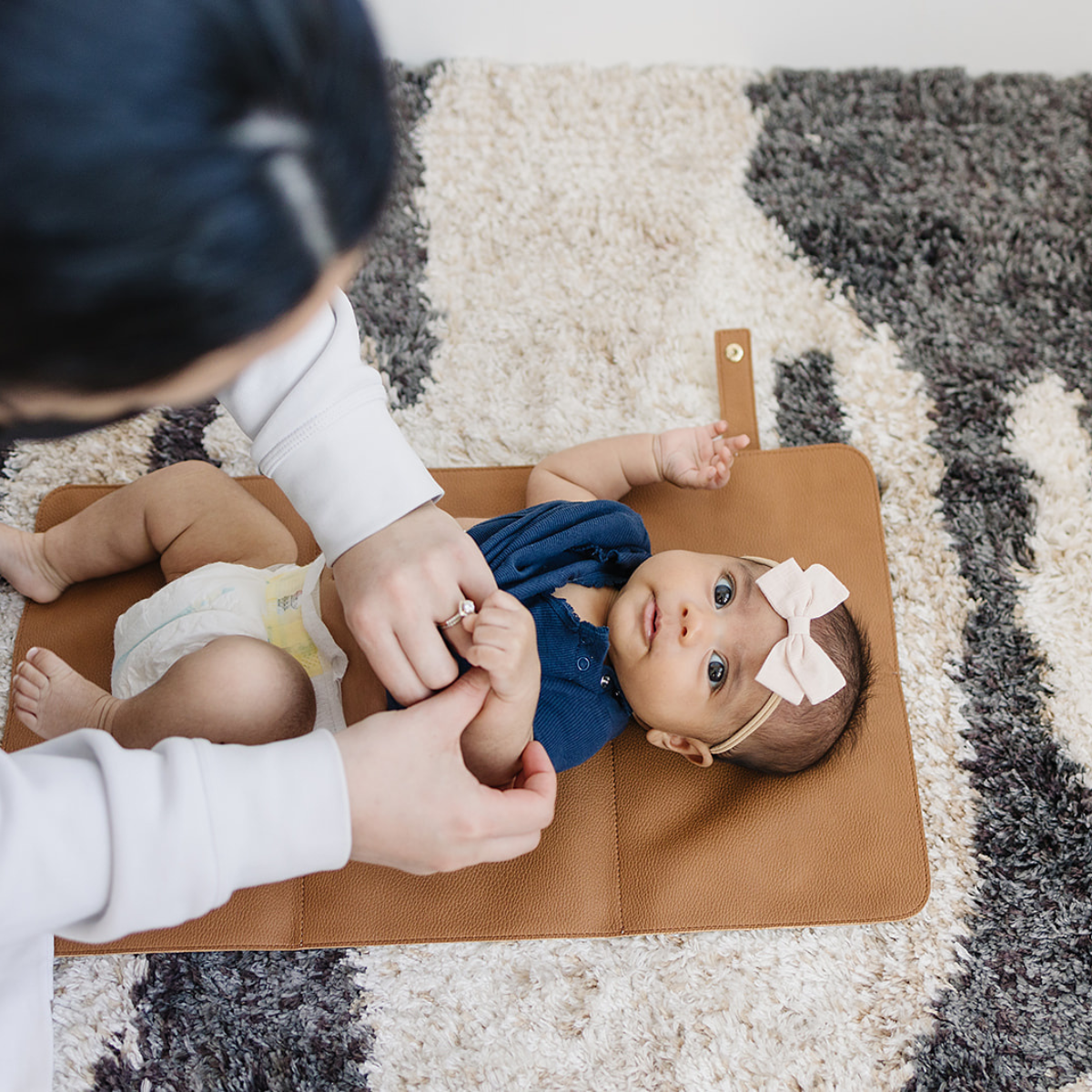 A caregiver holds a baby's hands as the little one, dressed in a blue outfit with a pink bow, rests on a vegan leather Changing Mat placed on a patterned rug.