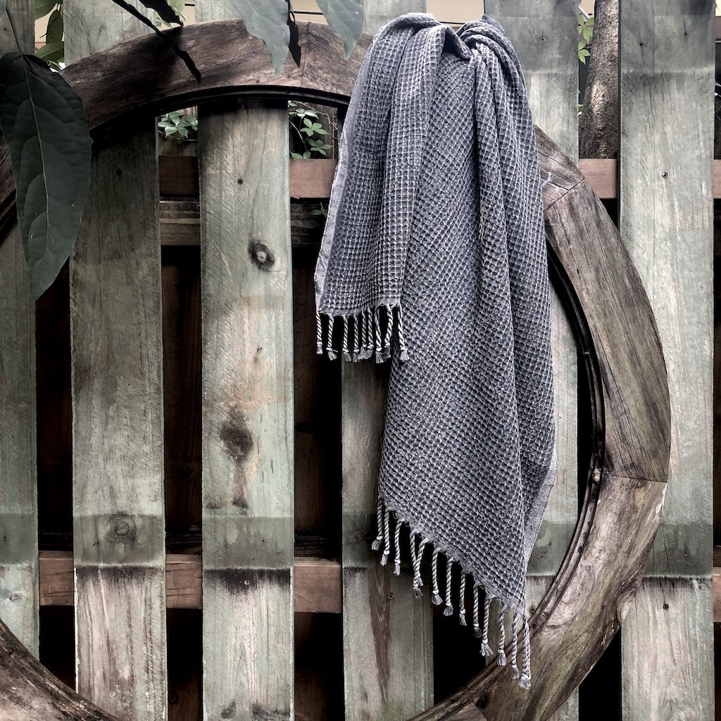 A folded, light gray blanket featuring a waffle texture and fringe tassels at the bottom mirrors the elegance of the Waffle Weave Turkish Towel, combining style and practicality for any cozy setting.