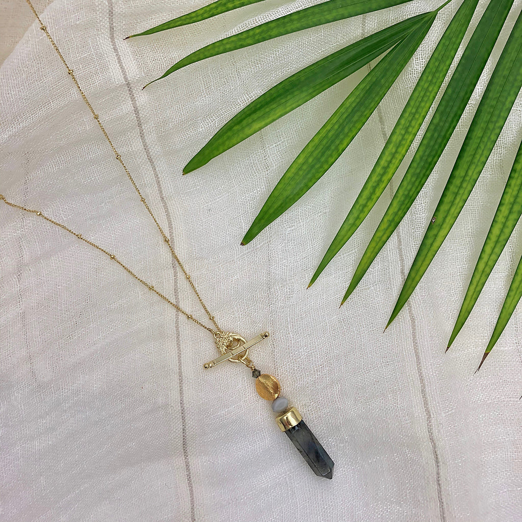 The Labradorite Lariat Necklace is a stunning gold necklace featuring a toggle clasp, a vertical rectangular labradorite stone pendant, and a round bead above it.