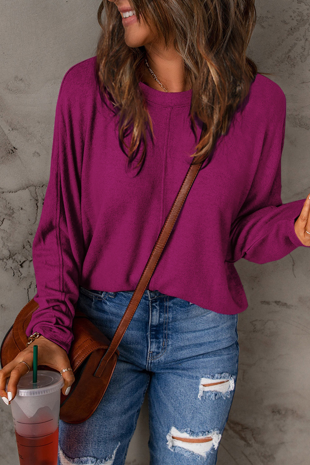 A person wearing a Double Take Full Size Round Neck Long Sleeve T-Shirt in green, blue distressed jeans with a slightly stretchy fit, and a crossbody bag, holding a beverage in a plastic cup.