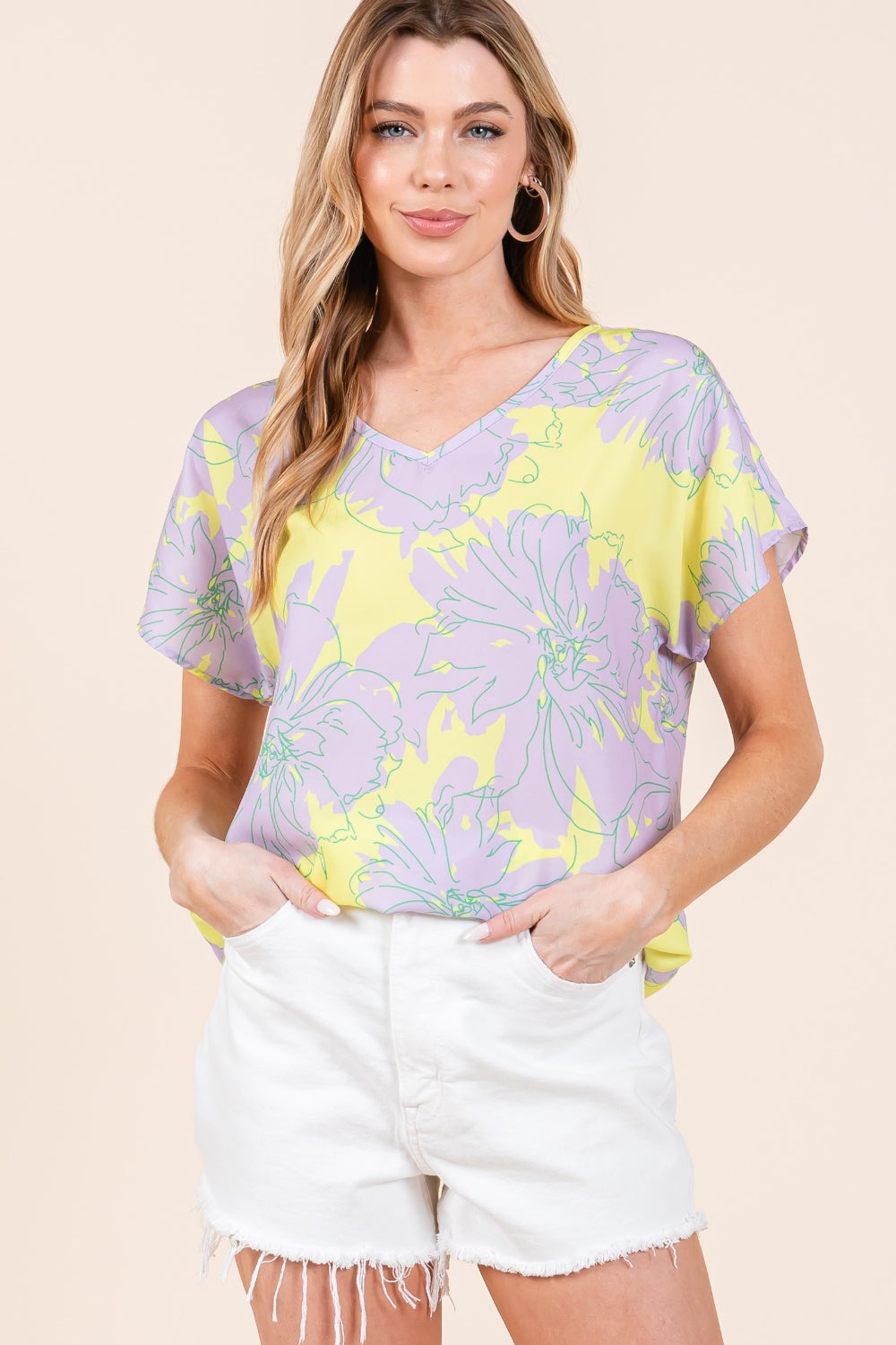 A woman wears the BOMBOM Floral Short Sleeve T-Shirt, featuring a vibrant floral print and a yellow V-neck, paired with white distressed shorts. She has long, wavy hair and stands against a beige background, showcasing her versatile and stylish look.