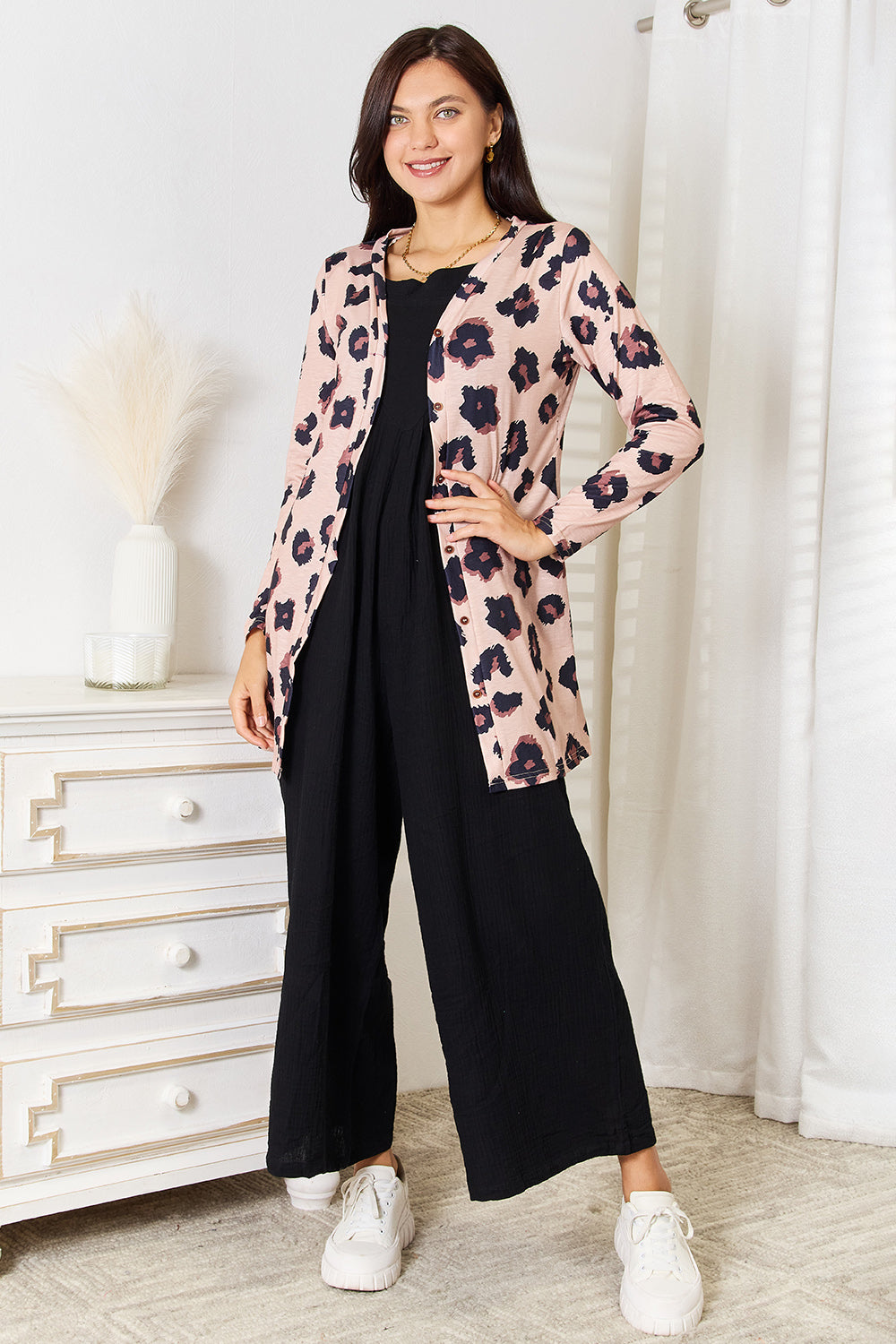 A woman wearing the Double Take Printed Button Front Longline Cardigan, featuring a pink floral pattern, over a black dress, stands indoors near a white vase with dried plants.