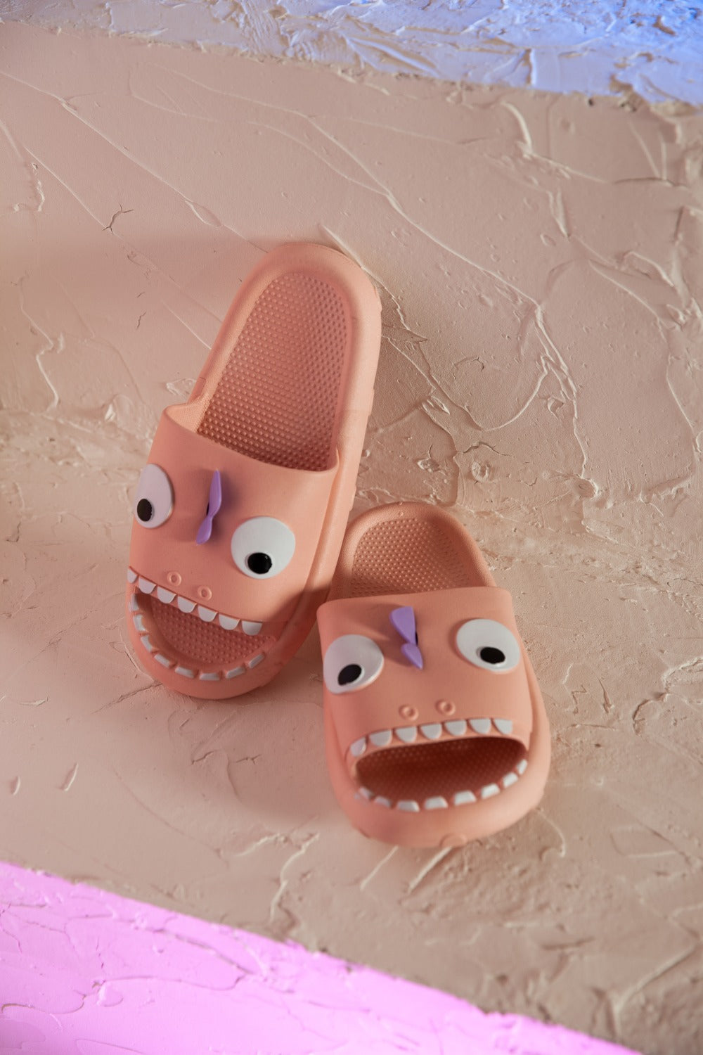 The NOOK JOI Monster Pillow Cloud Slides Non-Slip Slipper features cartoonish monster faces with large eyes, a purple nose, and an open mouth with teeth. Made of durable EVA material, these fun pink cloud slippers also boast an anti-slip design for added safety.