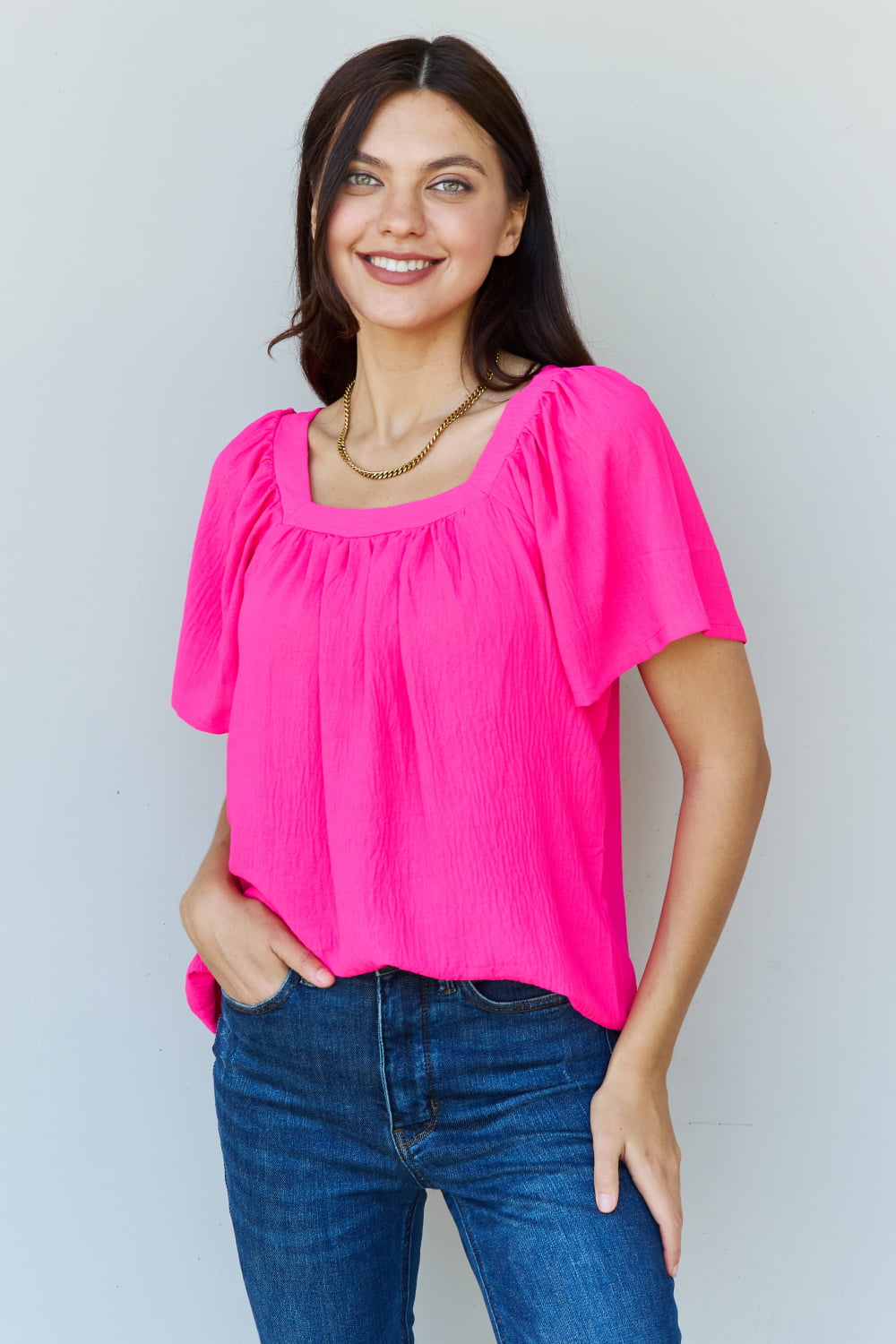 A person wearing the Ninexis Keep Me Close Square Neck Short Sleeve Blouse in Fuchsia paired with blue jeans smiles while posing against a plain background.