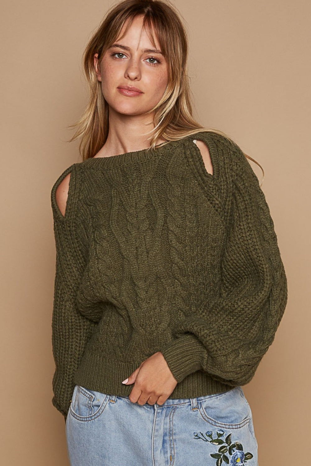 A woman wears the POL Cable Knit Cutout Long Sleeve Sweater in olive-green, showcasing a subtle shoulder keyhole, beautifully matched with light blue jeans adorned with floral embroidery, all set against a beige background.