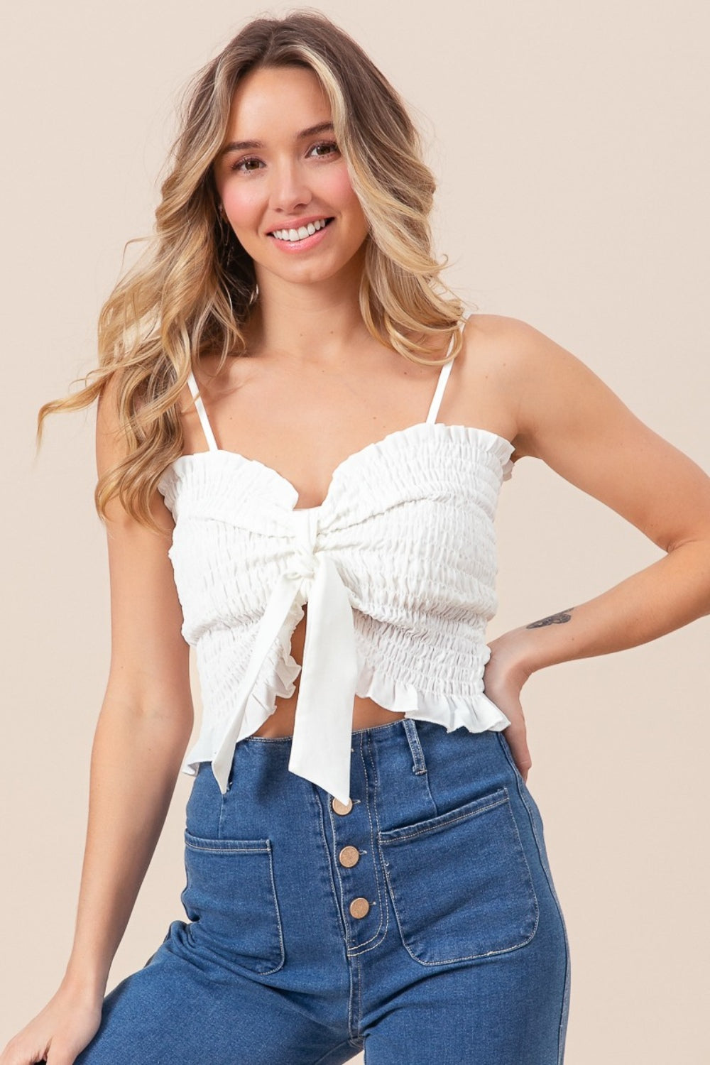 A woman wearing the BiBi Ruffled Smocked Ribbon Detail Cami in white, paired with blue high-waisted jeans, is posing against a neutral background.