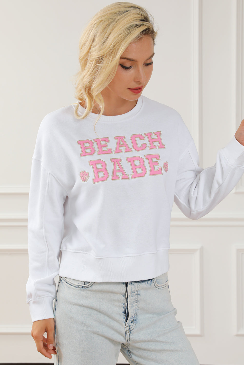 A person with long blonde hair is standing with their back to the camera, wearing a White BEACH BABE Slogan Graphic Casual Sweatshirt and light blue jeans.