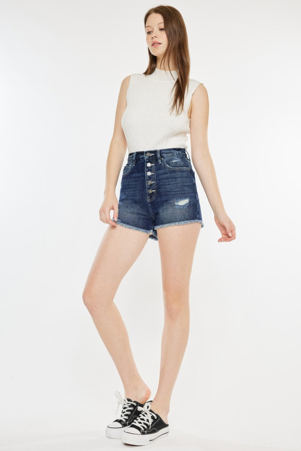 A woman stands against a plain background wearing a sleeveless white top, Kancan Raw Hem Button Fly Denim Shorts, and black slip-on sneakers.