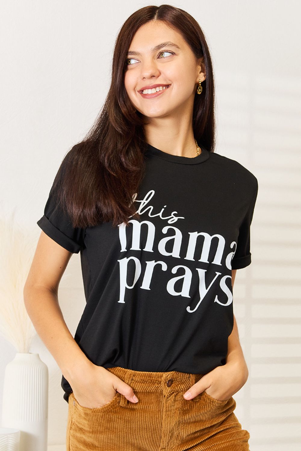 A woman wearing a Simply Love THIS MAMA PRAYS Graphic T-Shirt, paired with brown pants, stands gracefully in a softly lit room, exuding a sense of calm and spiritual gift.