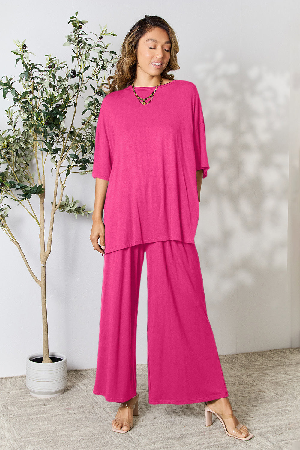 A woman stands near a potted plant and wooden ladder, wearing the Double Take Full Size Round Neck Slit Top and Pants Set in a bright pink two-piece outfit with matching wide-leg pants that are available in plus size and are easily machine washable.