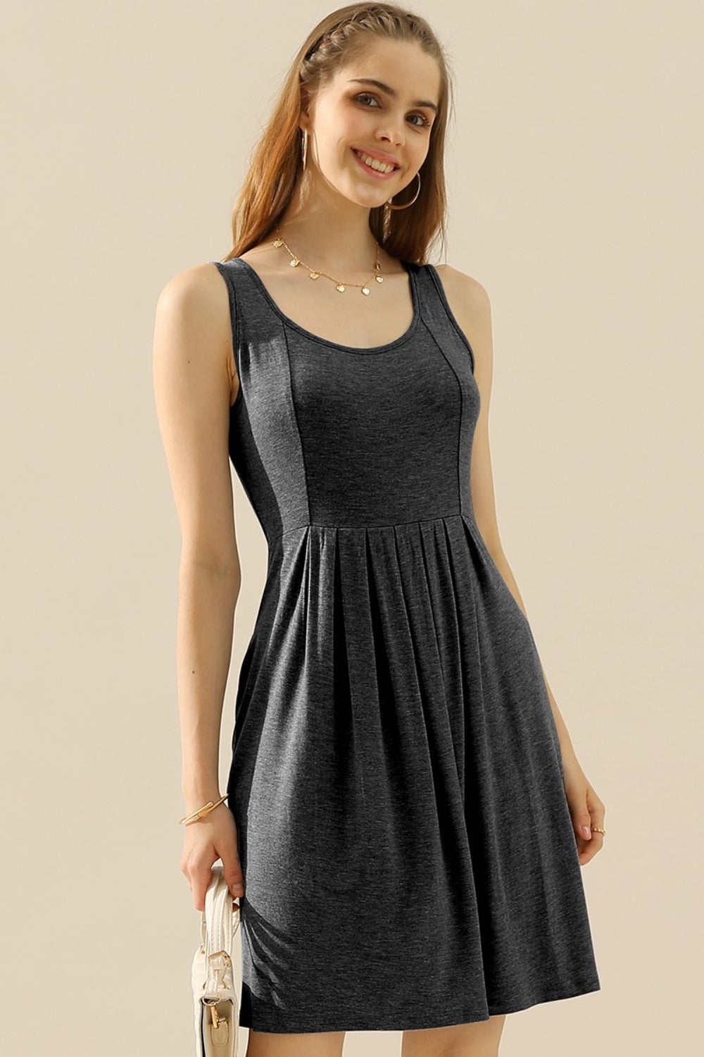 A woman in the Doublju Full Size Round Neck Ruched Sleeveless Dress with Pockets, featuring a stretchy gray fabric, smiles while holding a quilted white purse. She is wearing a gold necklace and bracelet.