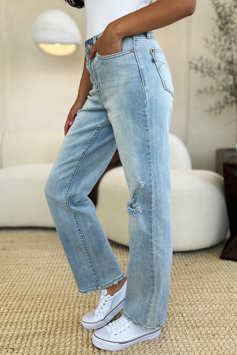 A person standing on a beige carpet, wearing Judy Blue Full Size High Waist Distressed Straight Jeans in light blue, a white top, and white sneakers.