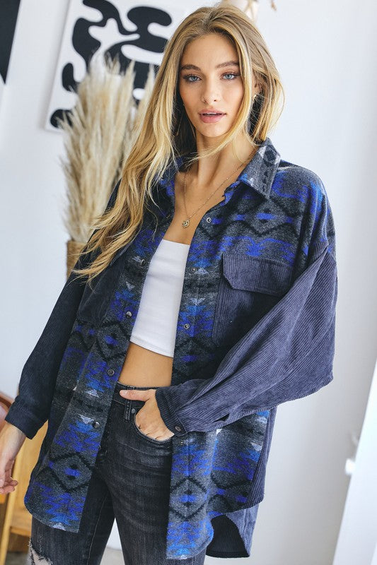 A woman with long blonde hair wears a blue and gray patterned shirt over a white crop top and dark jeans, standing indoors near a window, her casual **Printed Button Down Long Sleeve Jacket** draped casually over her shoulders.