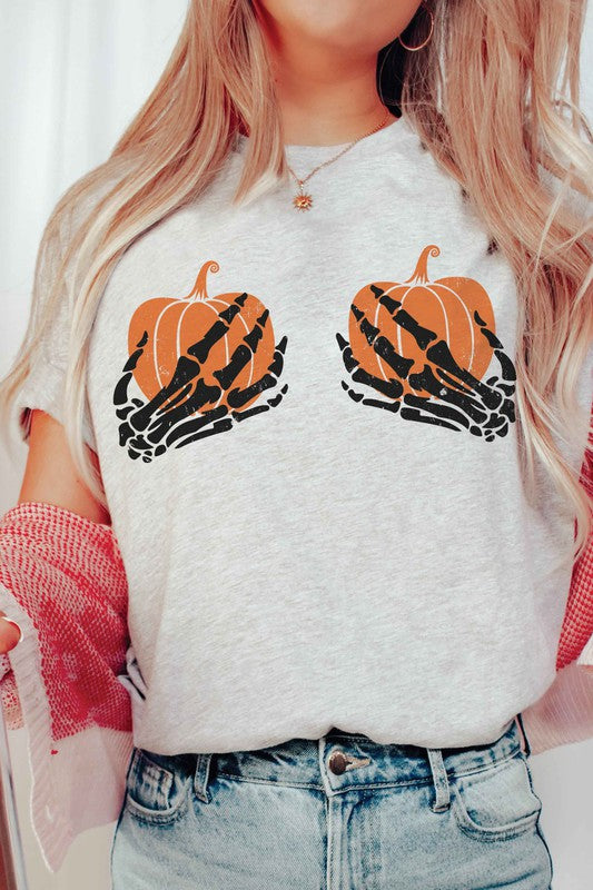 A person wearing the SKELETON HOLDING PUMPKINS Graphic Tee, which features two orange pumpkins and black skeletal hands, is sitting down. With long, wavy hair and accessorized with various bracelets and a necklace, the unisex sizing of the shirt makes it a versatile addition to any wardrobe.