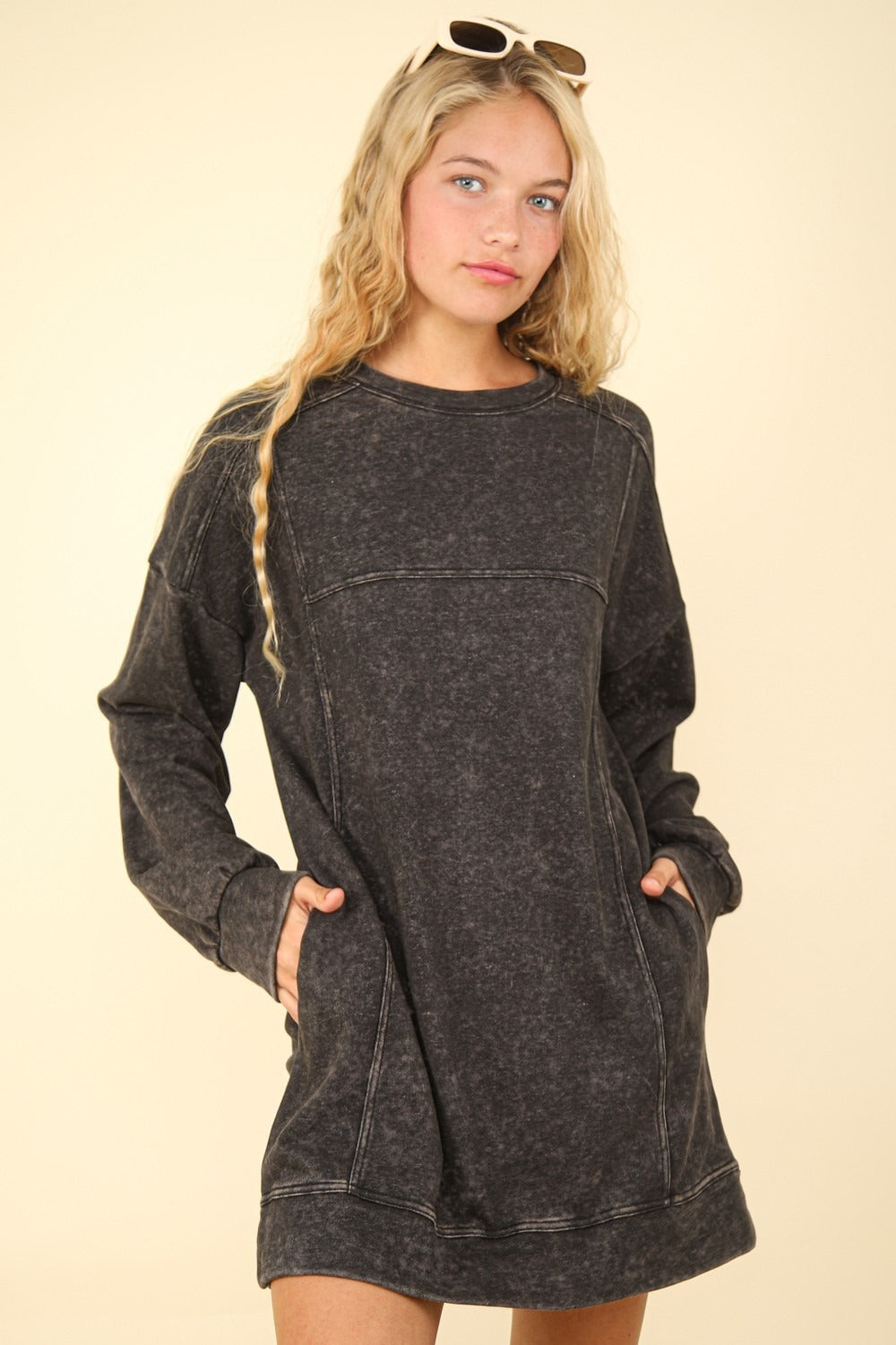 A person with long, wavy blonde hair wears a VERY J Mineral Washed Oversized Sweatshirt Mini Dress with pockets and sunglasses on top of their head, set against a beige background.