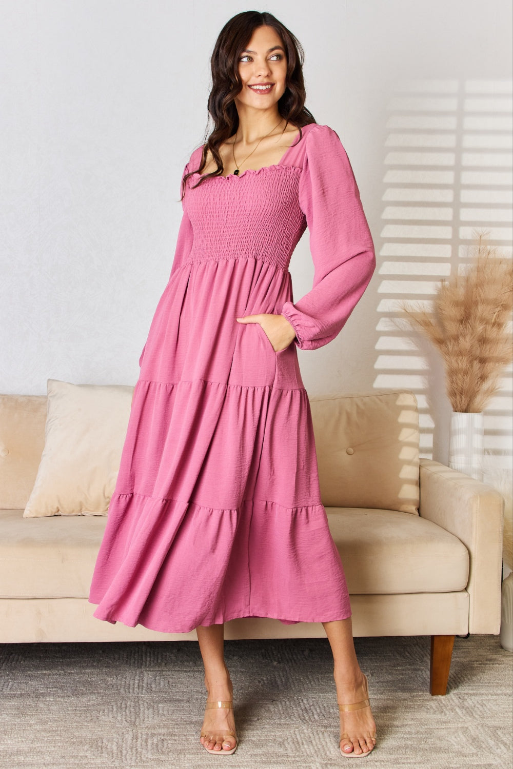 A woman in a Culture Code Full Size Ruffle Trim Smocked Tiered Dress stands in front of a beige sofa adorned with decorative pillows and framed by dried pampas grass, embodying a graceful silhouette perfect for elegant evening affairs.