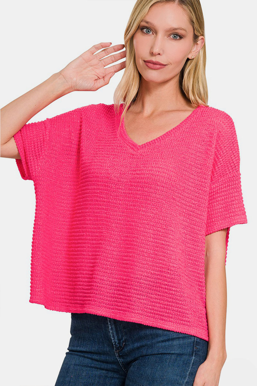 A woman wearing a loose-fitting, bright pink Zenana Drop Shoulder Short Sleeve Jacquard Knit Top with a V-neck and blue jeans touches her hair with one hand and looks at the camera.