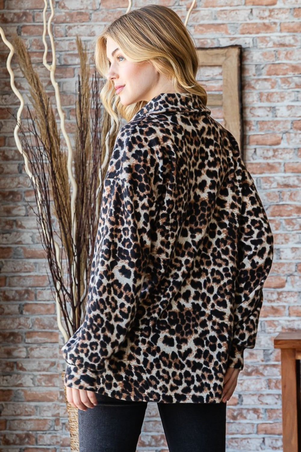 A person wearing a Heimish Full Size Leopard Johnny Collar Long Sleeve Top paired with black jeans stands in front of a brick wall adorned with wooden decor.